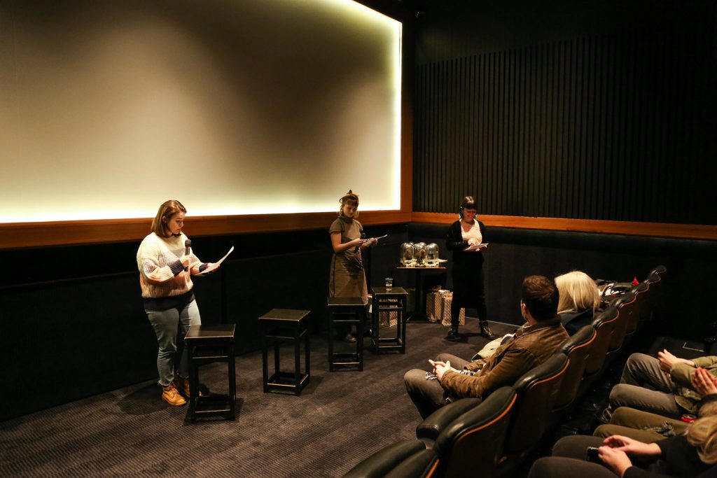A white young woman holding a microphone reads from a paper in front of a blank and lit cinema screen. To her right there are two other white young women and between them a table with trophies on it. In front of them there is a row of cinema seats and two white people seating on them.